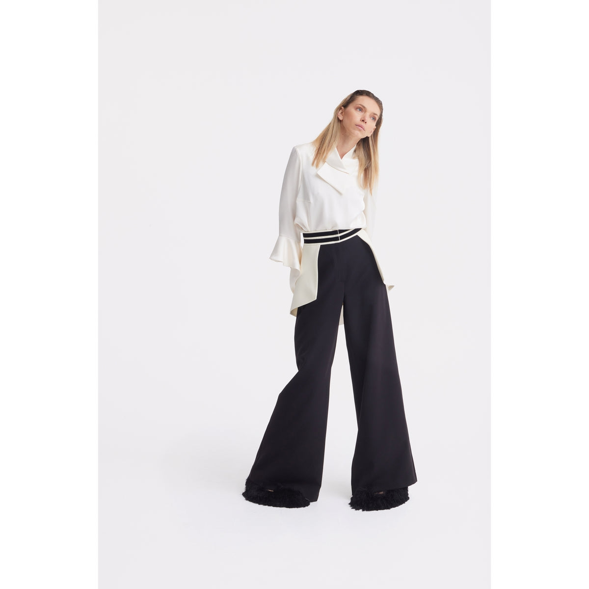 A woman stands against a white background, wearing a JULIA ALLERT Asymmetric Collar Satin Blouse in Ivory with flared sleeves and high-waisted, wide-leg black pants with a belt. She gazes slightly upward with a relaxed posture.