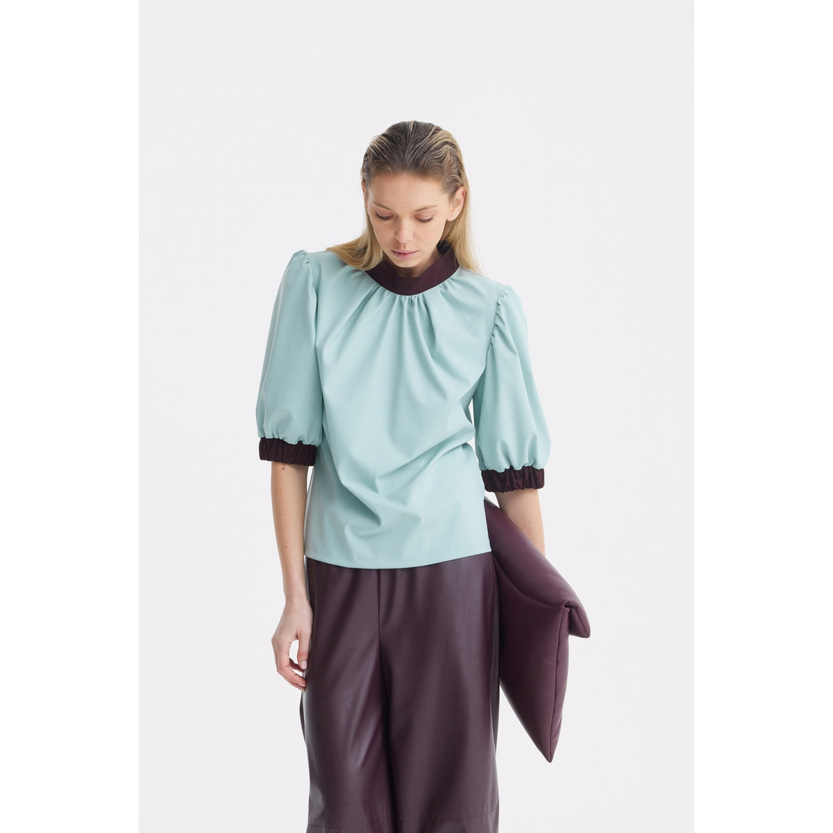 A person in a JULIA ALLERT Textured Short Sleeve Blouse in light blue, paired with dark high-waisted pants, stands against a plain white background holding a large dark-colored clutch.