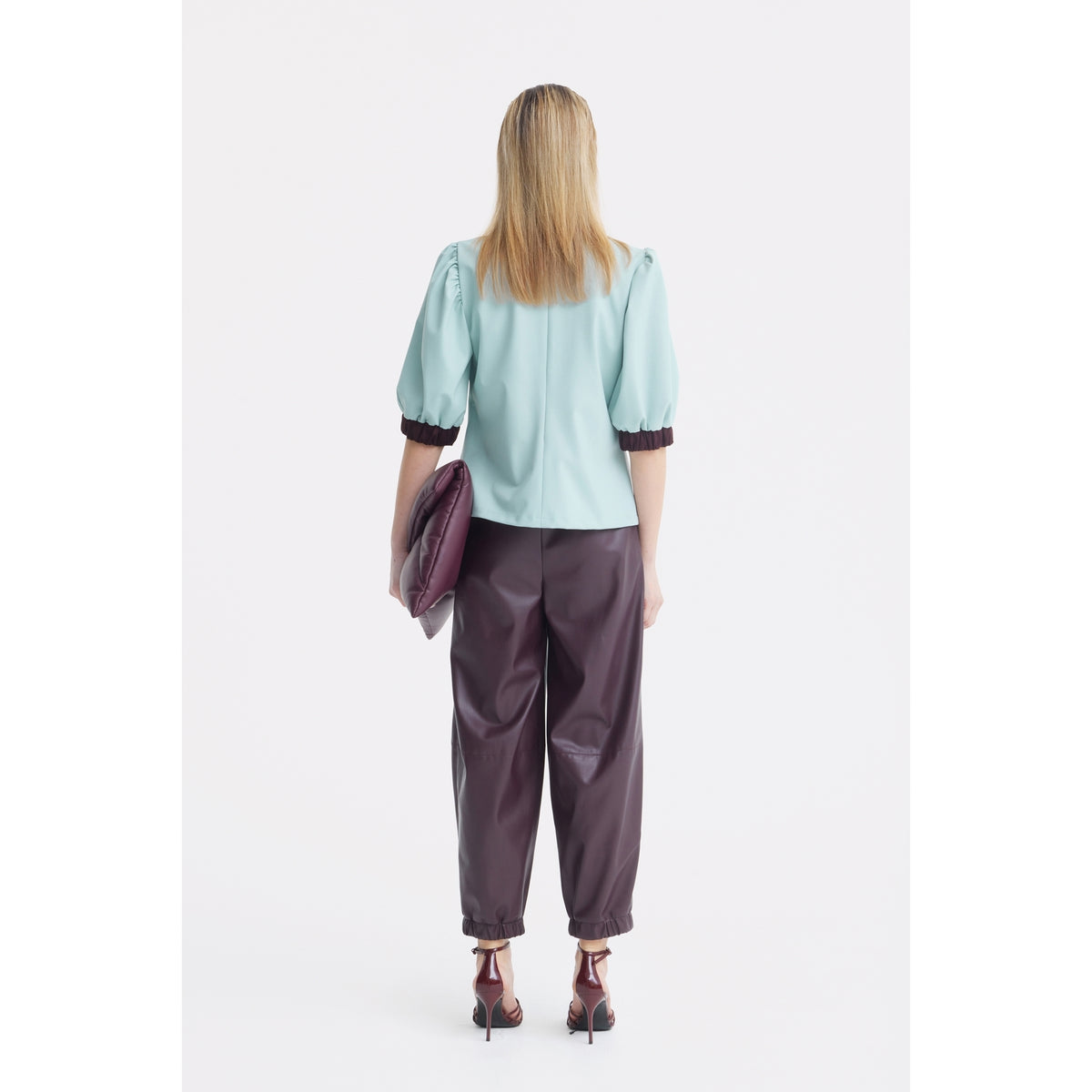 A woman stands facing away, wearing a JULIA ALLERT Textured Short Sleeve Blouse in light blue, paired with loose dark brown pants and maroon heels. She holds a large maroon clutch bag in her left hand.