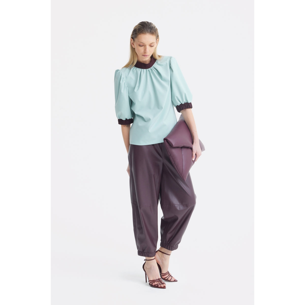 A woman poses against a plain white background wearing the JULIA ALLERT Textured Short Sleeve Blouse in light blue, styled with loose dark pants, strappy heels, and holding a large folded purple clutch.