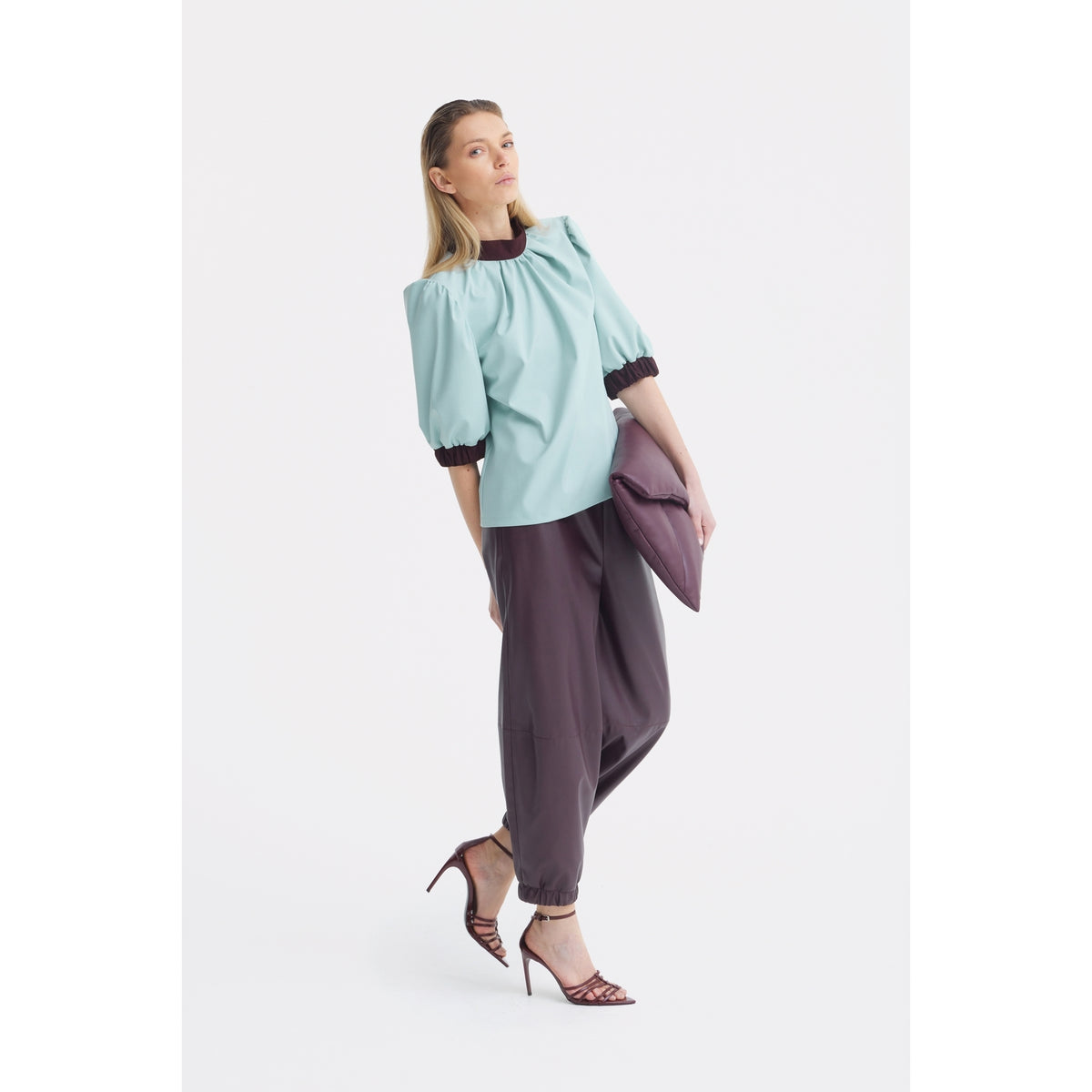 A woman stands against a white background wearing the JULIA ALLERT Textured Short Sleeve Blouse in light blue, paired with loose dark gray pants and strappy high heels. She holds a large purple clutch and gazes slightly upward.