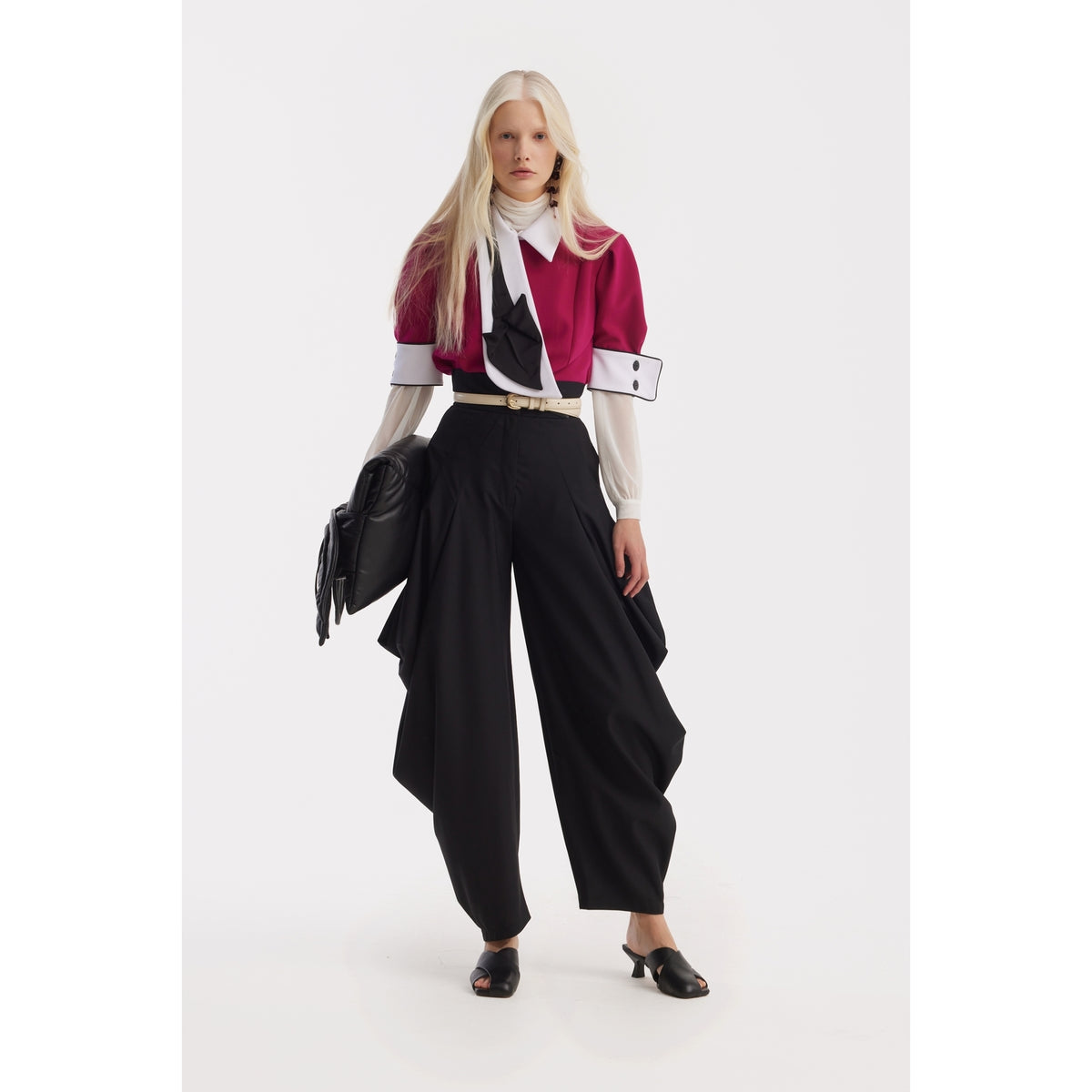 A model with platinum blonde hair poses in an avant-garde outfit featuring a magenta and white jacket over a sheer top, Couture Draped Wide-Leg Pants in black by JULIA ALLERT, black mules, and a sleek black clutch.
