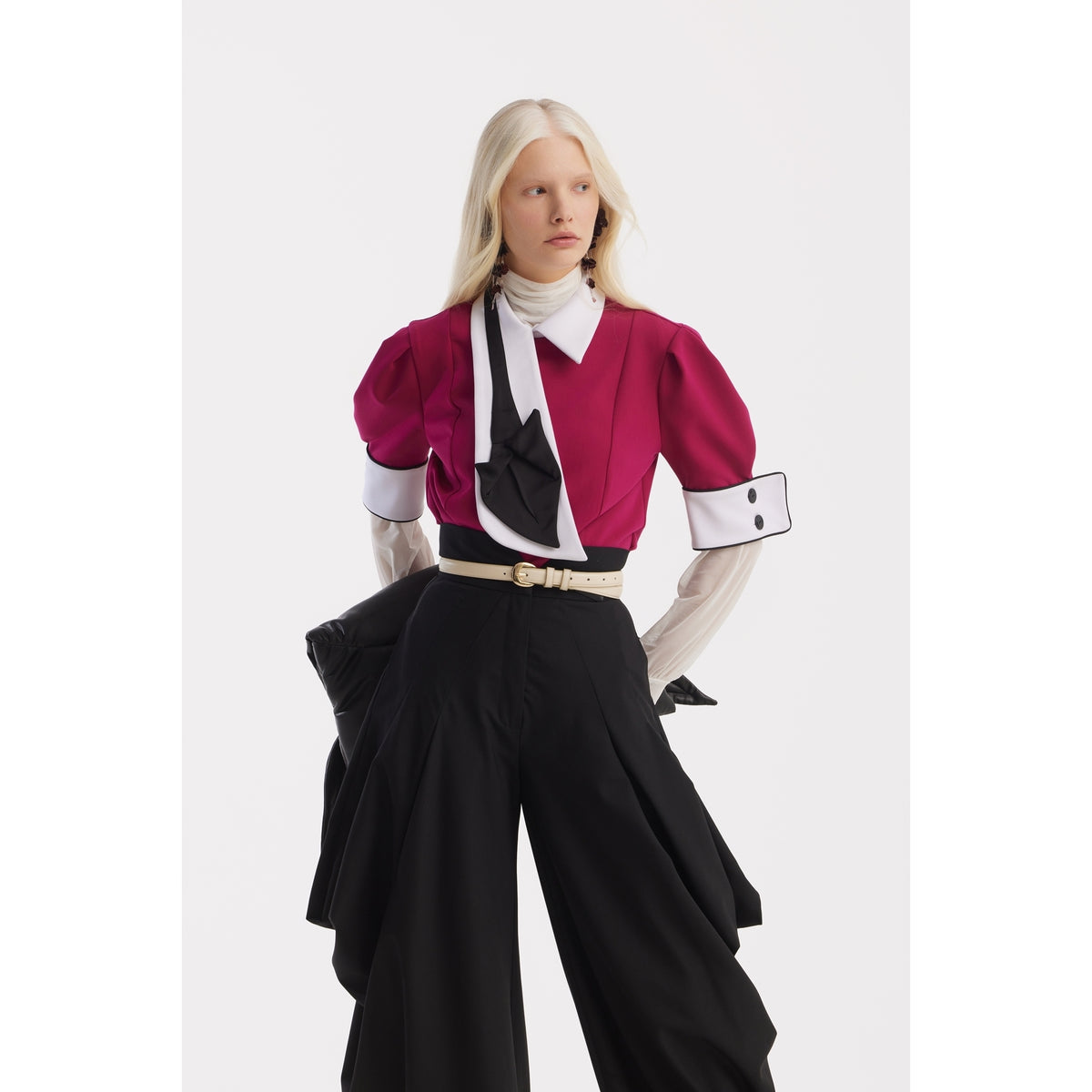 A person with long, straight blonde hair wears a magenta top with puffed sleeves, a white collar, and black-and-white accents, paired with JULIA ALLERT Couture Draped Wide-Leg Pants in black and a beige-buckled belt, posing against a plain white background.