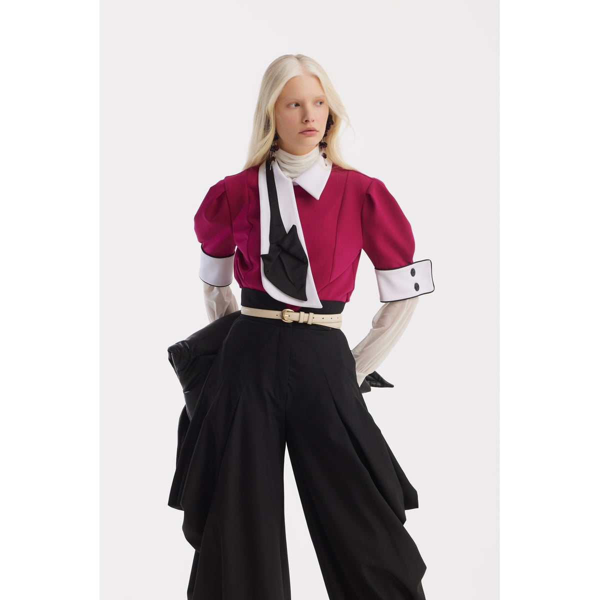 A woman with long platinum blonde hair models the JULIA ALLERT Statement Blouse With White Collar And Cuffs - Fuchsia, styled with a black necktie, wide-leg black pants, and gloves. She poses against a plain white background, gazing to the side.