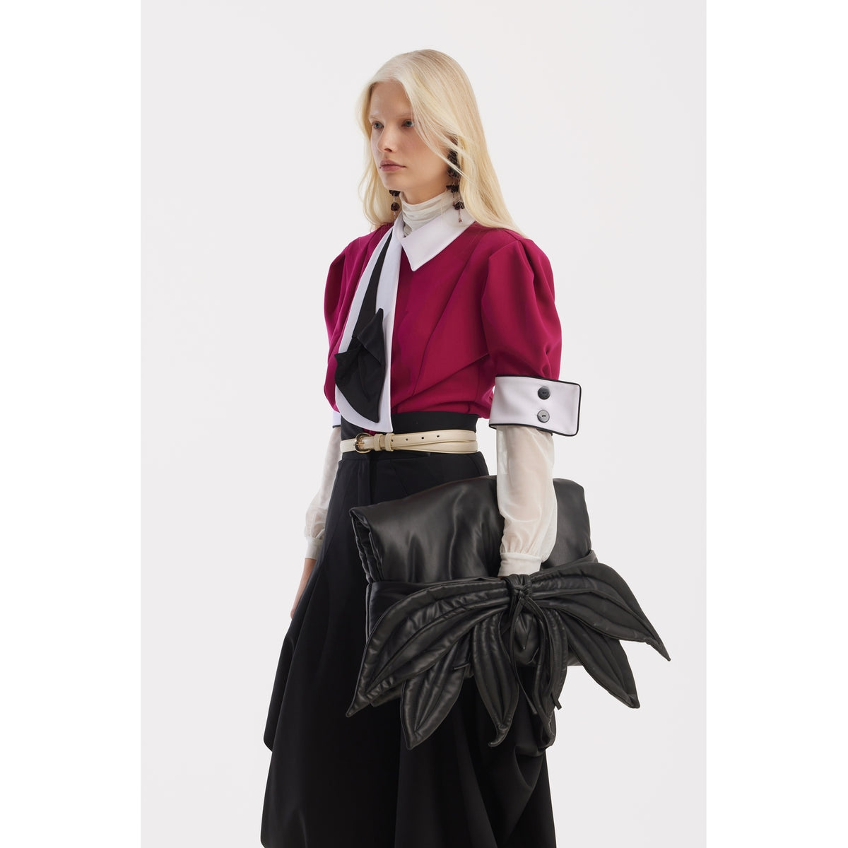 A person with long blond hair wears a JULIA ALLERT Statement Blouse With White Collar And Cuffs in fuchsia, styled with a black and white necktie, cream belt, long black skirt, and carries a large black leaf-shaped bag.