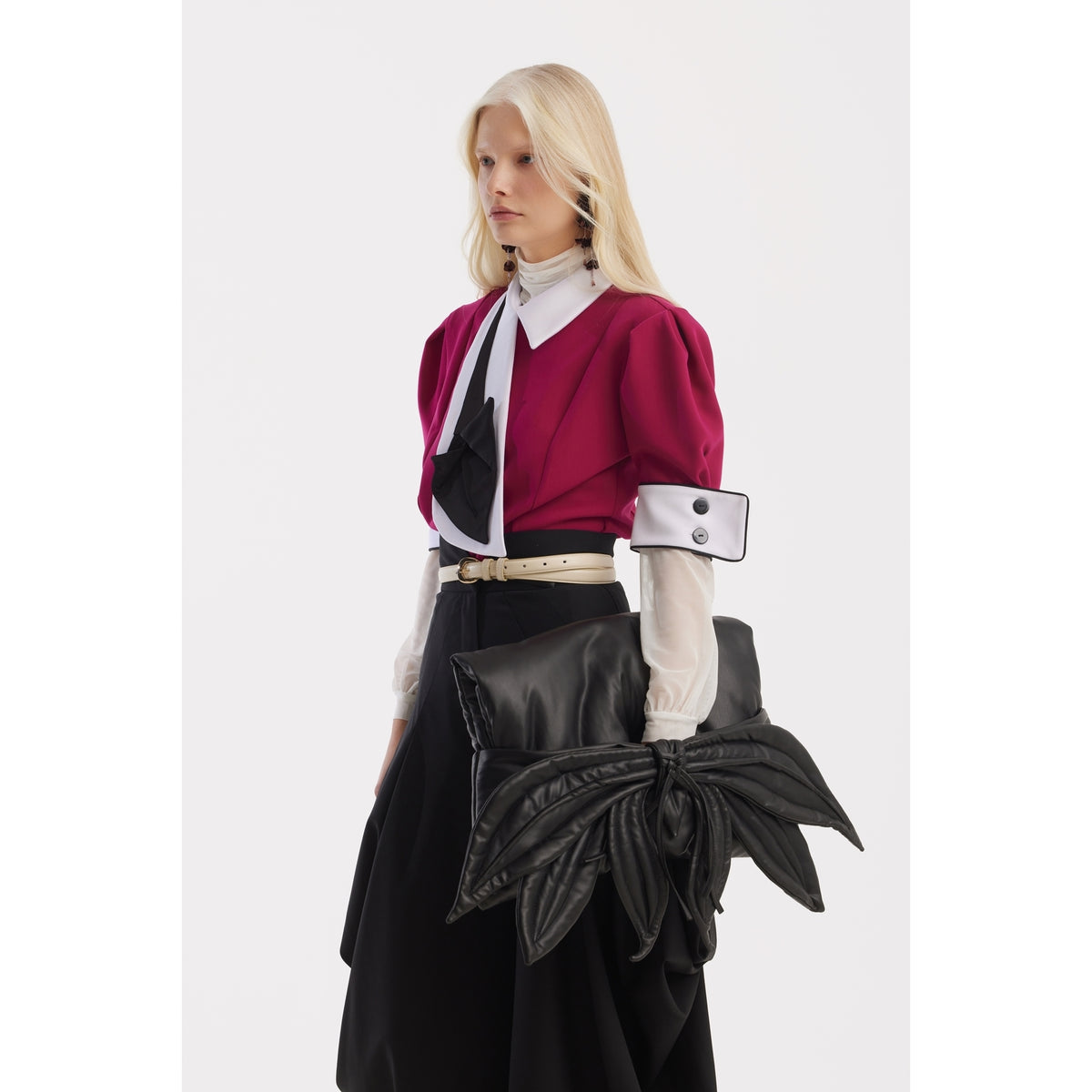 A woman with long blonde hair wears a magenta jacket, a white blouse with black accents, and JULIA ALLERT Couture Draped Wide-Leg Pants in black. She carries an oversized sculptural clutch shaped like large leaves.