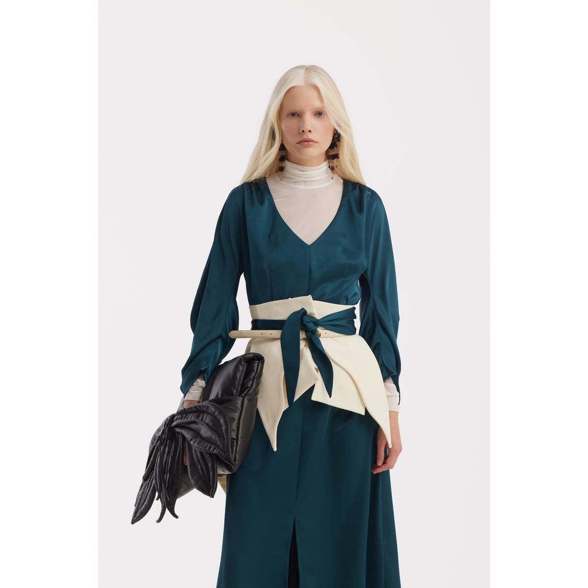A woman with long blonde hair wears a JULIA ALLERT Satin Ankle-Length Dress With Belt in turquoise, layered over a white turtleneck. She accessorizes with statement earrings, a large black glove-shaped purse, and a wide cream bow belt.