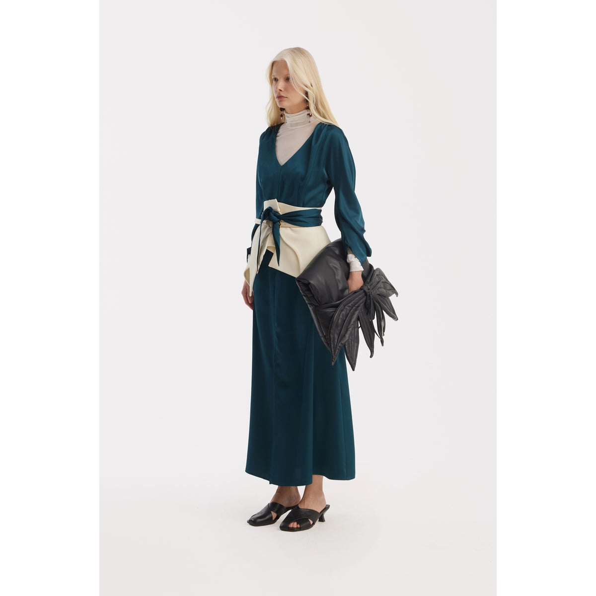 A woman in a JULIA ALLERT Satin Ankle-Length Dress With Belt in turquoise, layered over a light gray turtleneck, holds a large black spiked clutch and wears black open-toe heels against a white background.