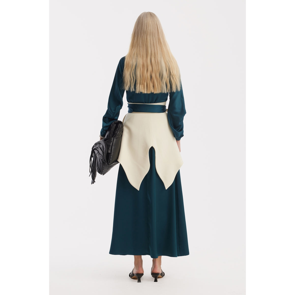 A person with long, straight blonde hair faces away, wearing the JULIA ALLERT Satin Ankle-Length Dress With Belt in turquoise and holding a black bag, paired with heeled shoes against a plain white background.