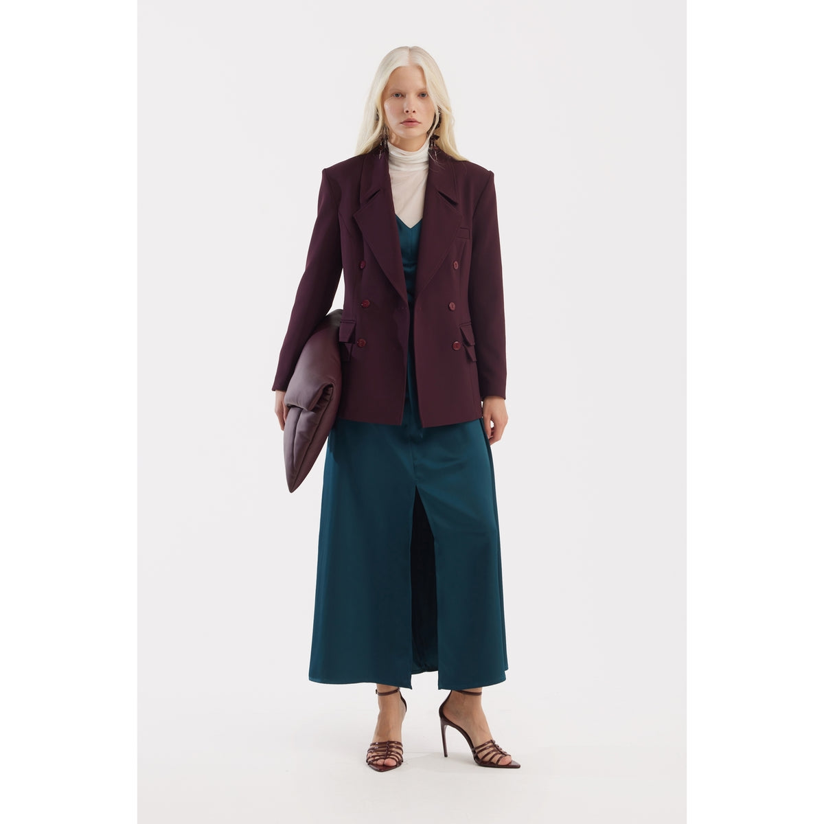 A woman with long blonde hair wears the JULIA ALLERT Satin Ankle-Length Dress With Belt in turquoise under a burgundy blazer, paired with strappy heels and a large clutch, set against a plain white background.