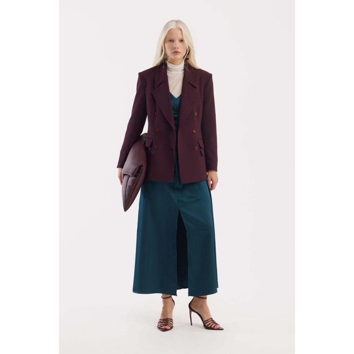 A woman with long blonde hair wears the JULIA ALLERT Classic Tailored Double-Breasted Blazer in burgundy over a teal dress, paired with a white turtleneck, strappy heels, and a matching clutch against a plain white background.