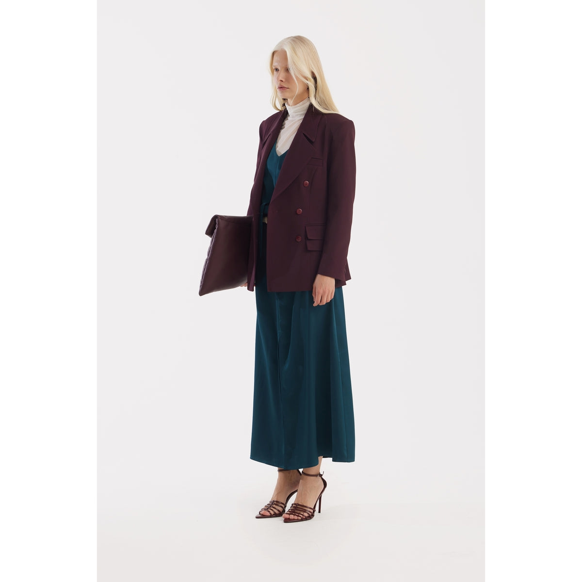 A woman with long blonde hair models the JULIA ALLERT Satin Ankle-Length Dress With Belt in turquoise, paired with a burgundy blazer, white turtleneck, strappy heels, and a matching clutch against a plain white background.