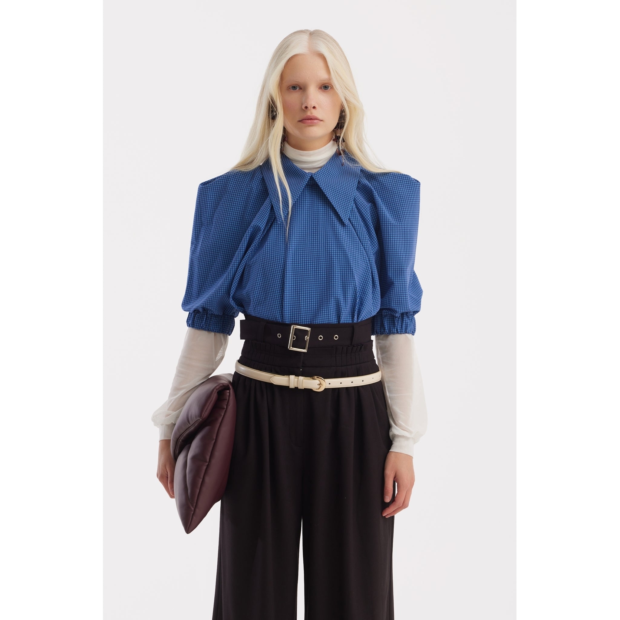 A person with long, straight, platinum-blonde hair wears a JULIA ALLERT Designer Plaid Puff Sleeve Blouse - Blue over a white turtleneck, high-waisted black belted pants, and holds a large brown clutch.