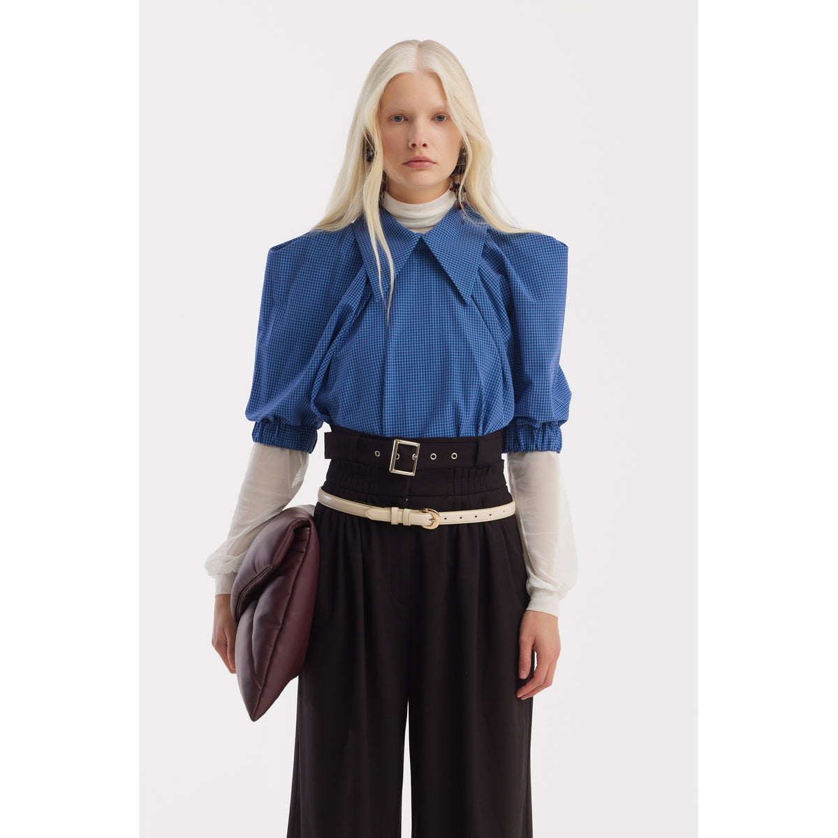 A person with long, straight, platinum-blonde hair wears a JULIA ALLERT Designer Plaid Puff Sleeve Blouse - Blue over a white turtleneck, high-waisted black belted pants, and holds a large brown clutch.