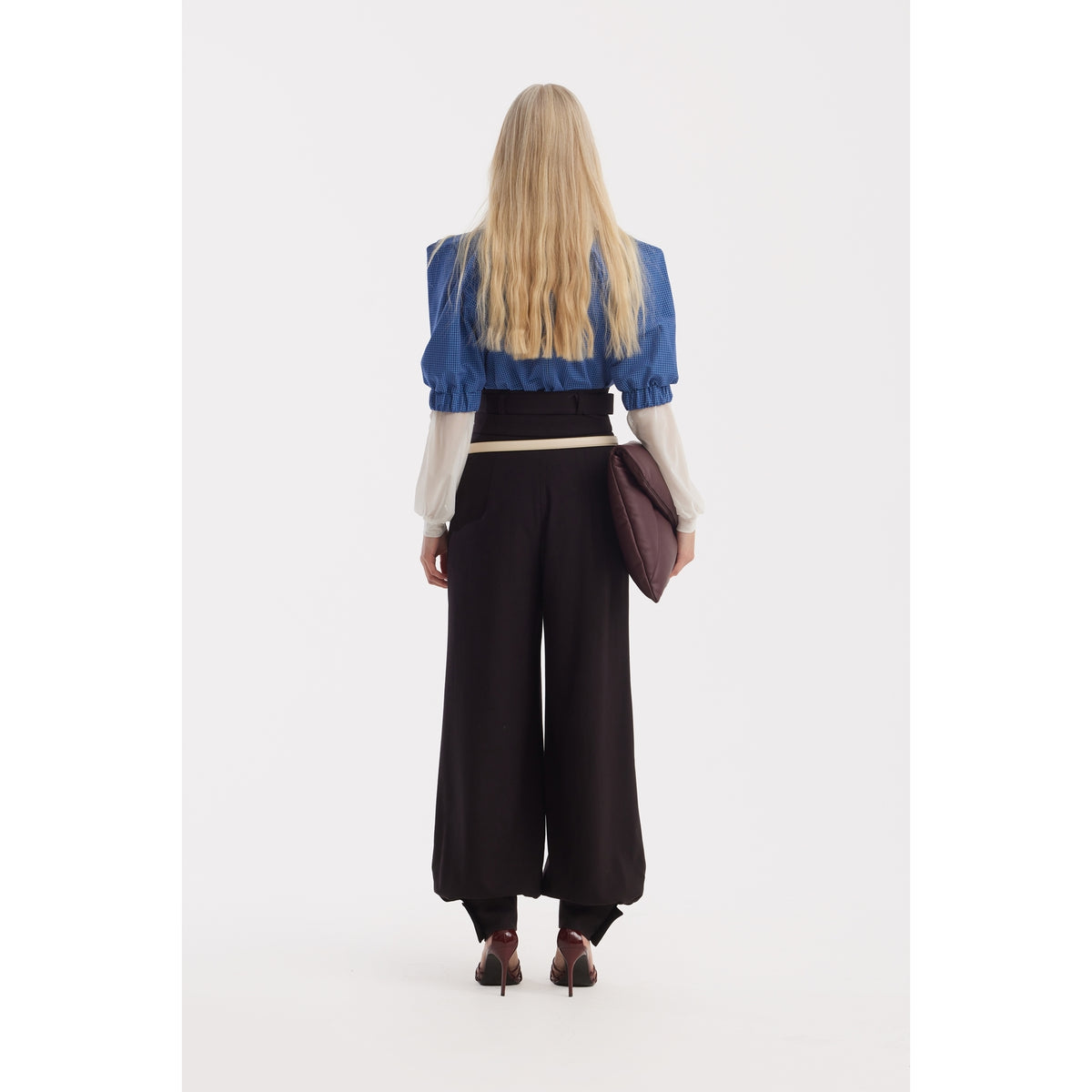 A woman with long blonde hair stands facing away, wearing a blue jacket with puffed sleeves, JULIA ALLERT Belted High-Waist Balloon-Leg Pants in black, maroon heels, and holding a dark brown clutch against a white background.