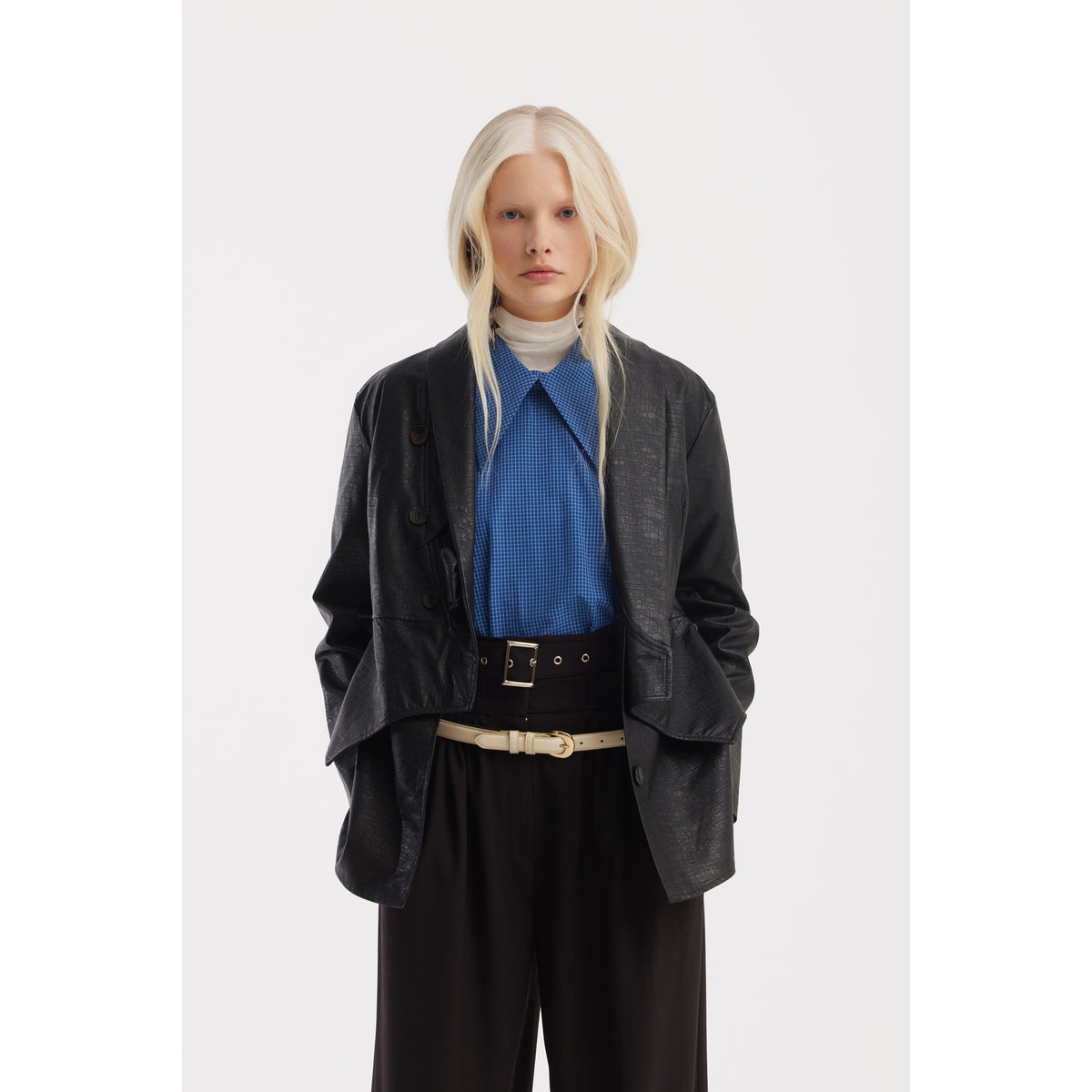 A person with long, straight blonde hair stands against a white background, wearing a black oversized jacket, blue shirt, white turtleneck, and JULIA ALLERT Belted High-Waist Balloon-Leg Pants in black.
