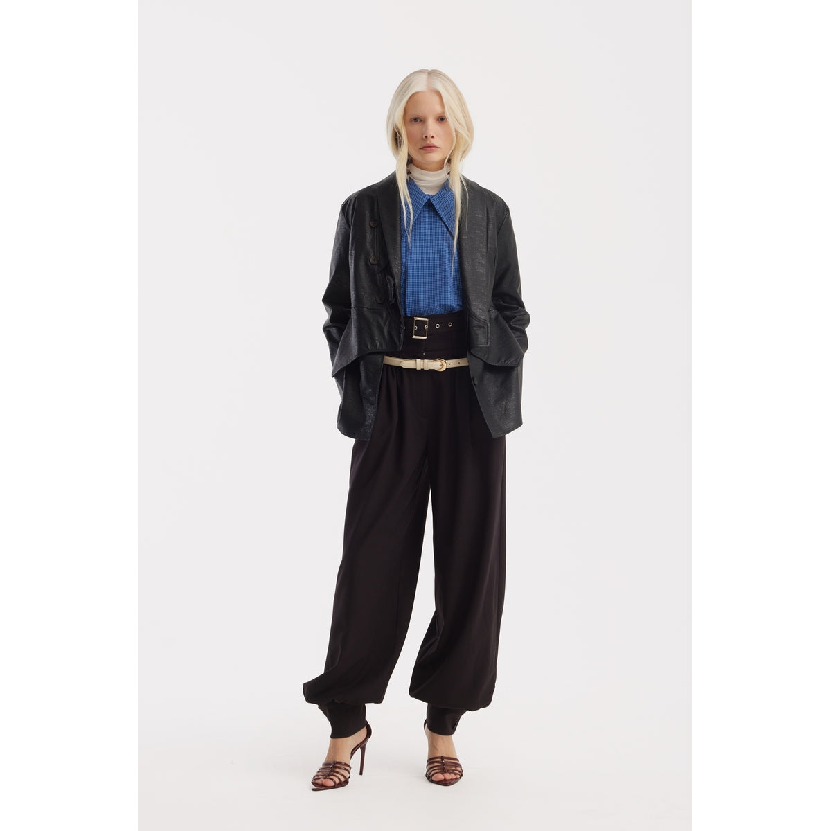 A woman with long blonde hair wears a JULIA ALLERT black leather jacket, Designer Plaid Puff Sleeve Blouse - Blue, white turtleneck, wide black trousers with a black belt, and brown heeled sandals, standing against a plain white background.