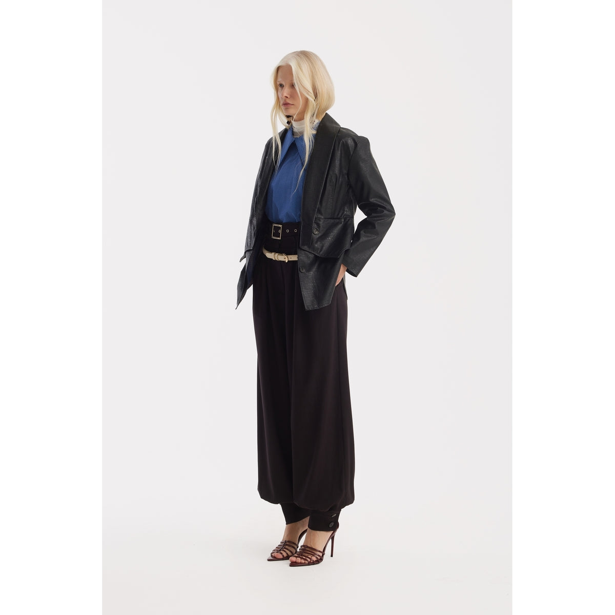 A person with long blonde hair stands against a white background, wearing a black leather jacket over the JULIA ALLERT Designer Plaid Puff Sleeve Blouse in blue, paired with black wide-leg pants and brown heeled sandals.