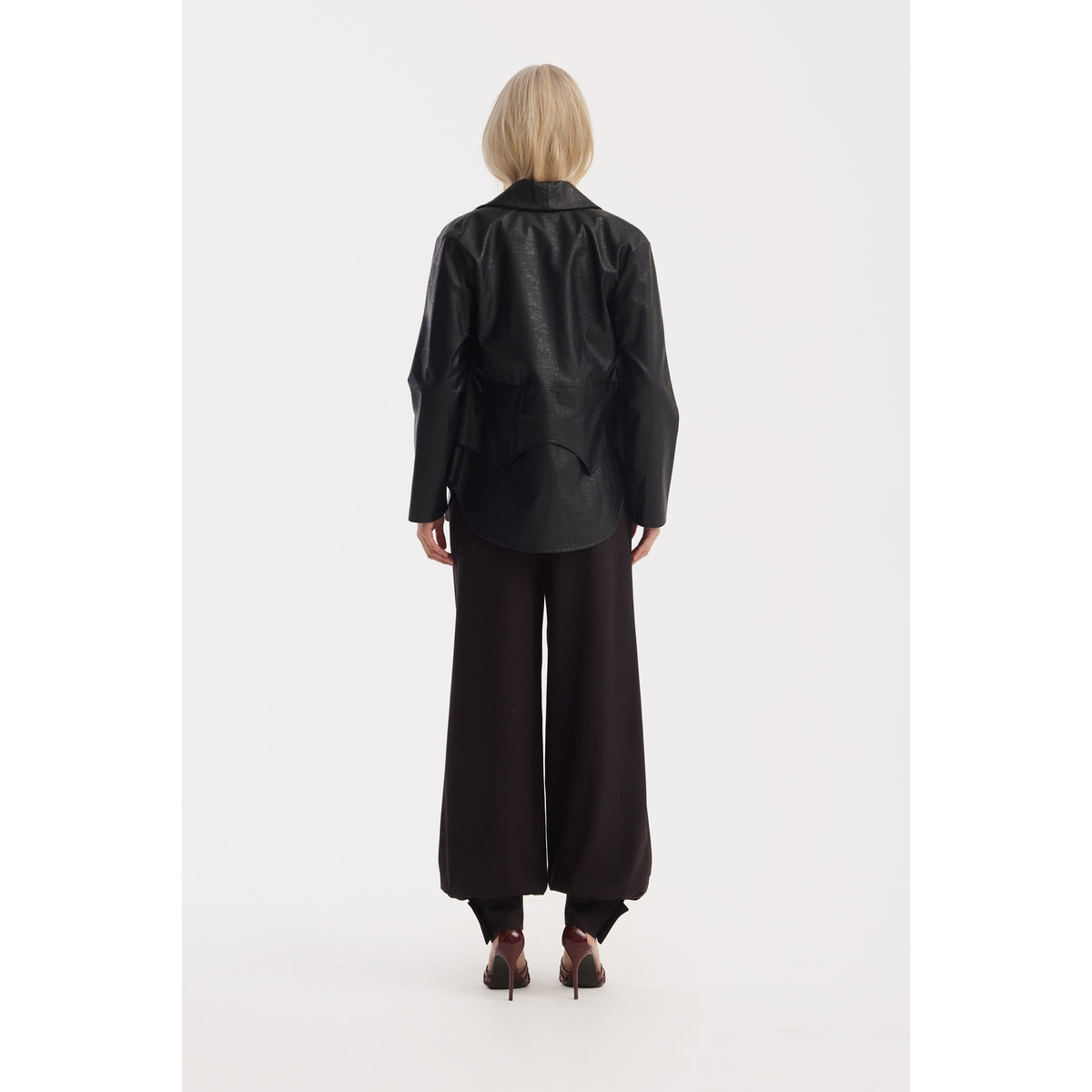 A person with short blonde hair stands facing away, wearing a loose black leather jacket, JULIA ALLERT Belted High-Waist Balloon-Leg Pants in black, and high-heeled shoes against a plain white background.