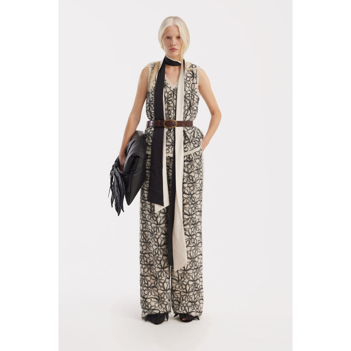 A person with straight blonde hair wears JULIA ALLERT's Elastic Waist Abstract Textured Wide Pants in ivory, a sleeveless patterned jumpsuit, wide brown belt, and black scarf. They hold a black jacket and stand against a plain white background.