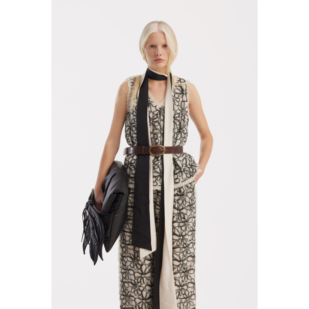 A person with light blond hair wears the JULIA ALLERT Sleeveless Abstract Textured Top in Ivory and matching wide-leg pants, accessorized with a black and beige scarf, brown belt, holding a black jacket against a plain white background.