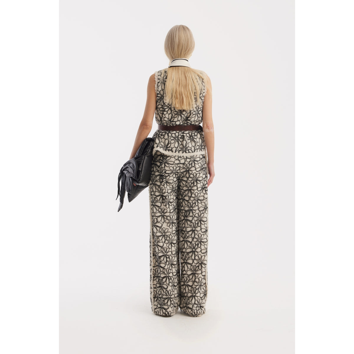 A woman with long blonde hair stands facing away, wearing JULIA ALLERT's Elastic Waist Abstract Textured Wide Pants in ivory, and holding a large black fringed bag against a plain white background.