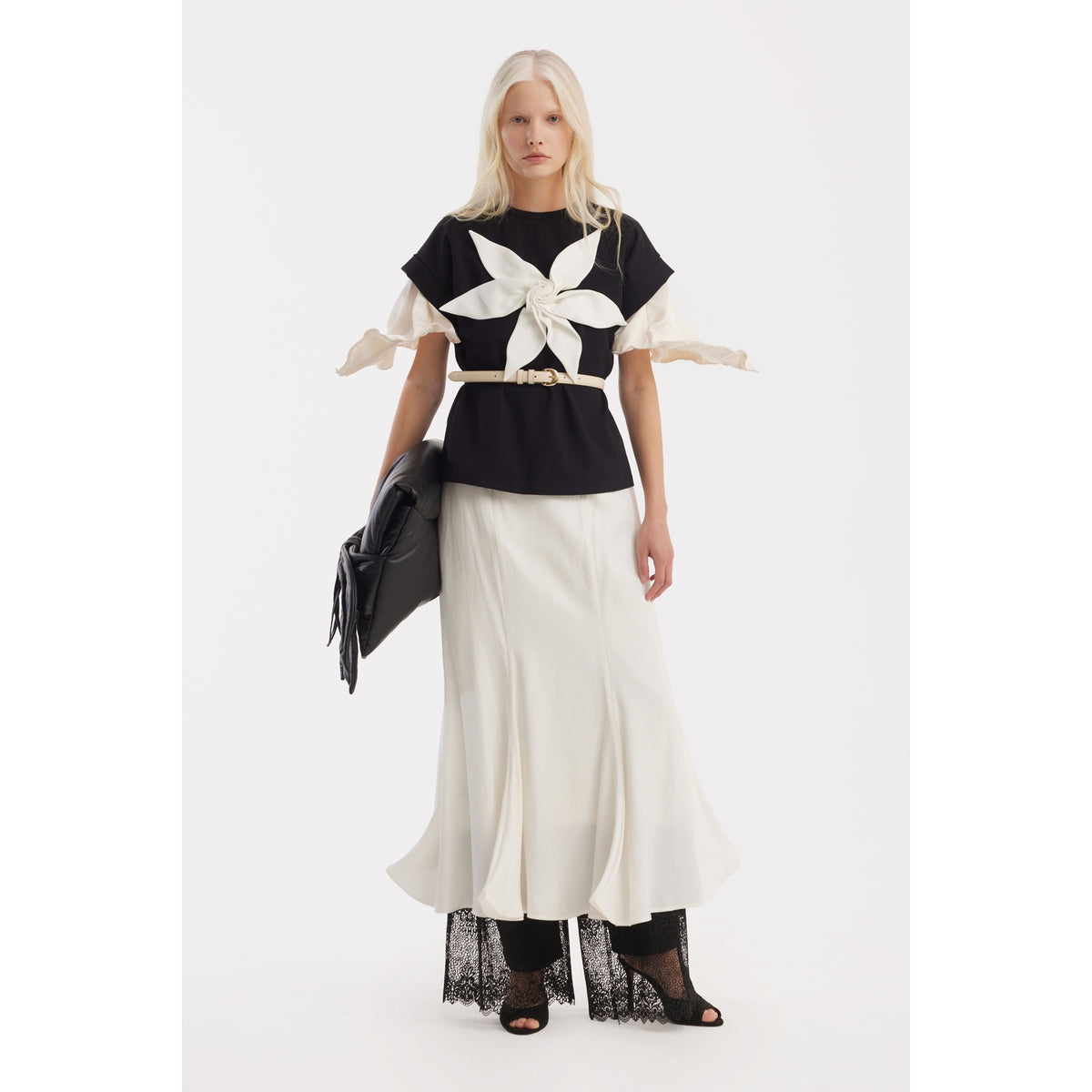 A woman with long blonde hair wears the JULIA ALLERT Crew-Neck Jersey T-Shirt With Flower in black, paired with a flowing white skirt, black lace pants, and black heels. She carries a large black bag and stands against a plain white background.