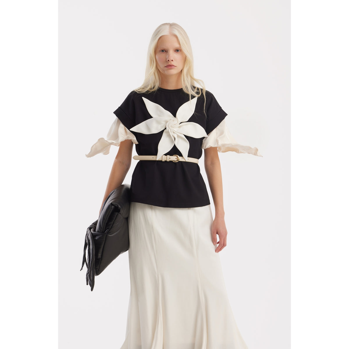 A woman with long blonde hair wears the JULIA ALLERT Crew-Neck Jersey T-Shirt With Flower in black, a cream skirt, and carries a black bag. The background is plain white.