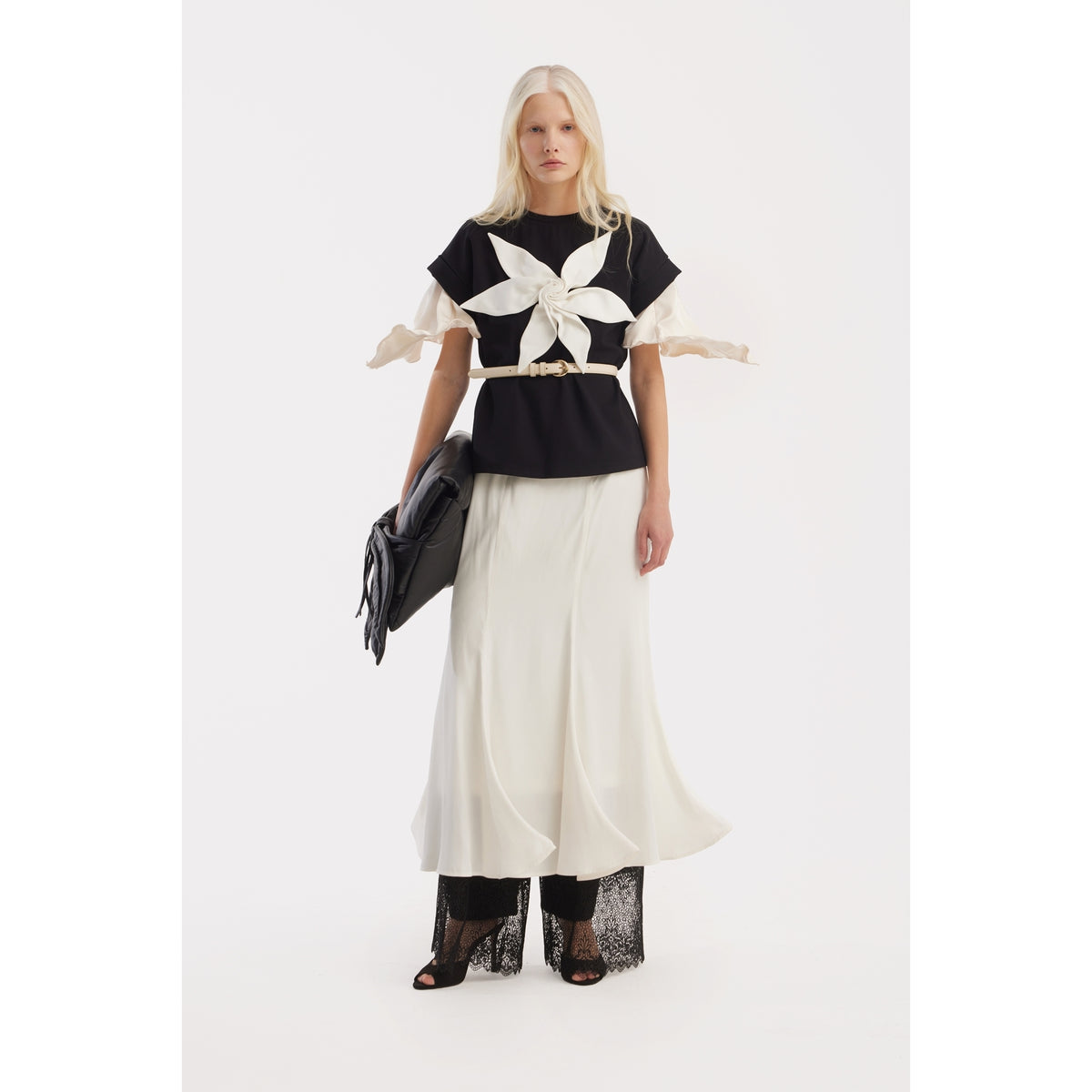 A woman with long blonde hair wears the JULIA ALLERT Crew-Neck Jersey T-Shirt With Flower in black, paired with a flowing white skirt, black lace pants, and open-toe heels, holding a black clutch against a plain white background.