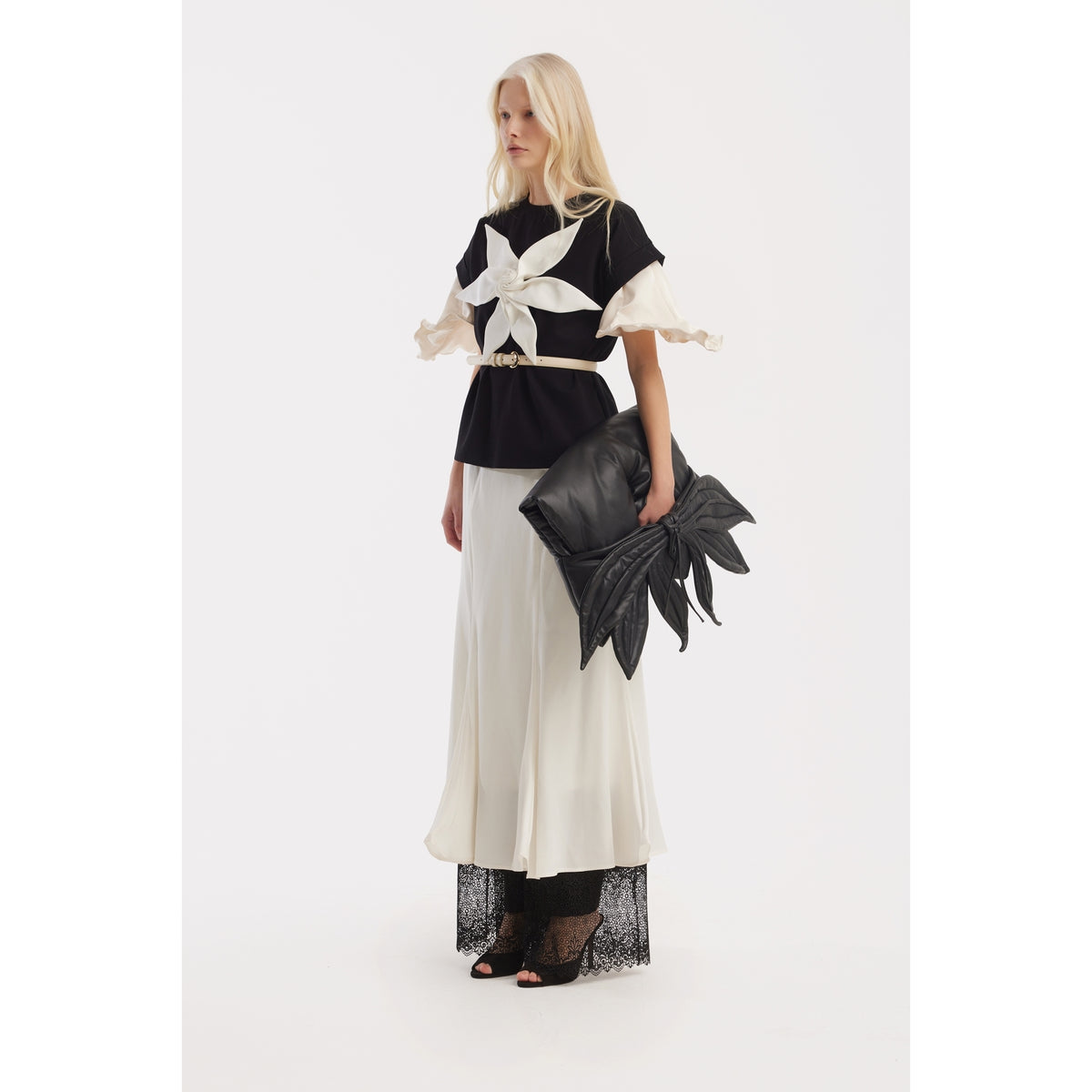 A woman with long blonde hair, wearing a JULIA ALLERT Crew-Neck Jersey T-Shirt With Flower in black, stands against a white background holding a black leaf-shaped clutch bag and wearing black heeled sandals.