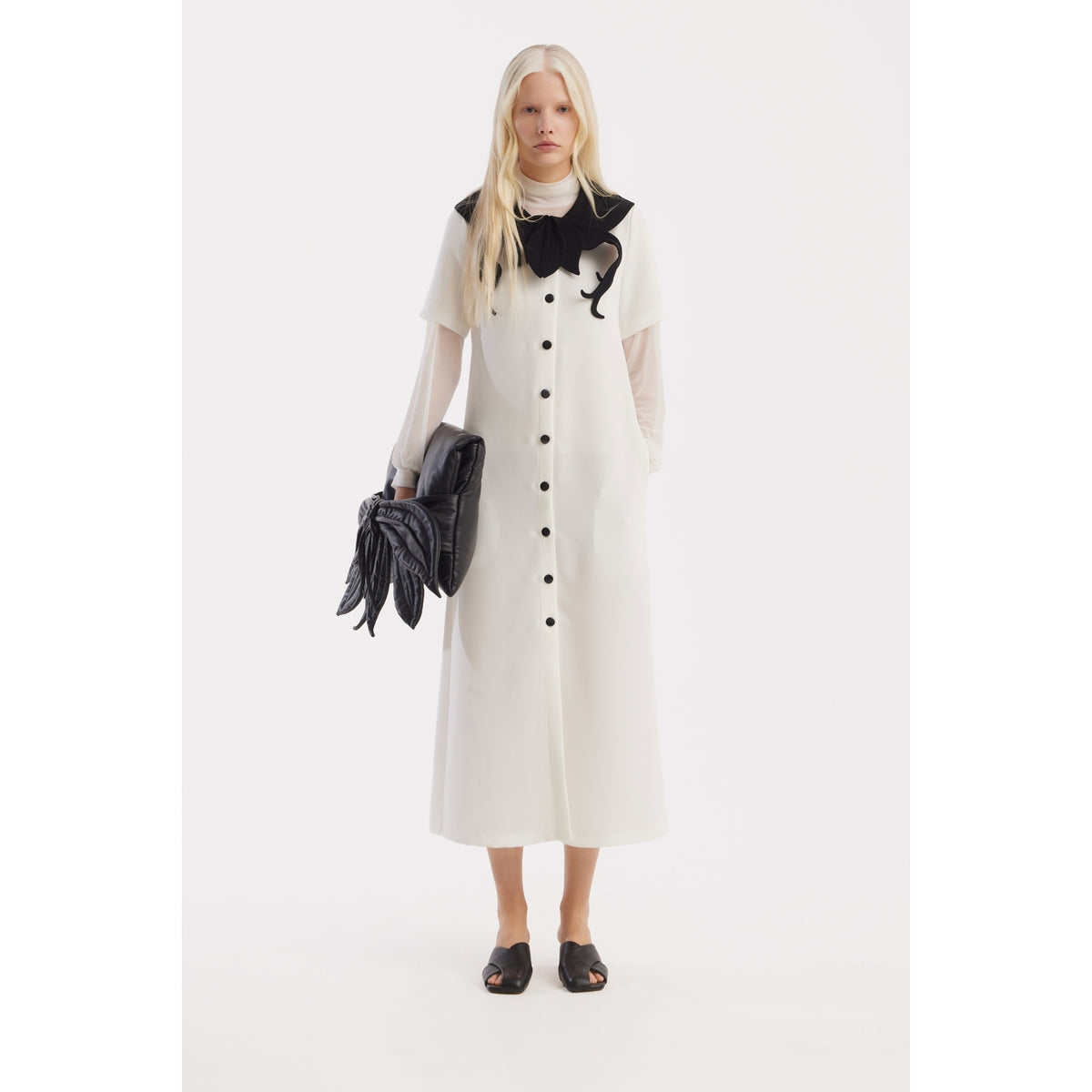 A woman with long blonde hair wears the JULIA ALLERT Straight Midi Shirt Dress With Adjustable Collar in white, featuring sheer sleeves and a large black bow. She holds a black glove-shaped clutch against a plain white background.