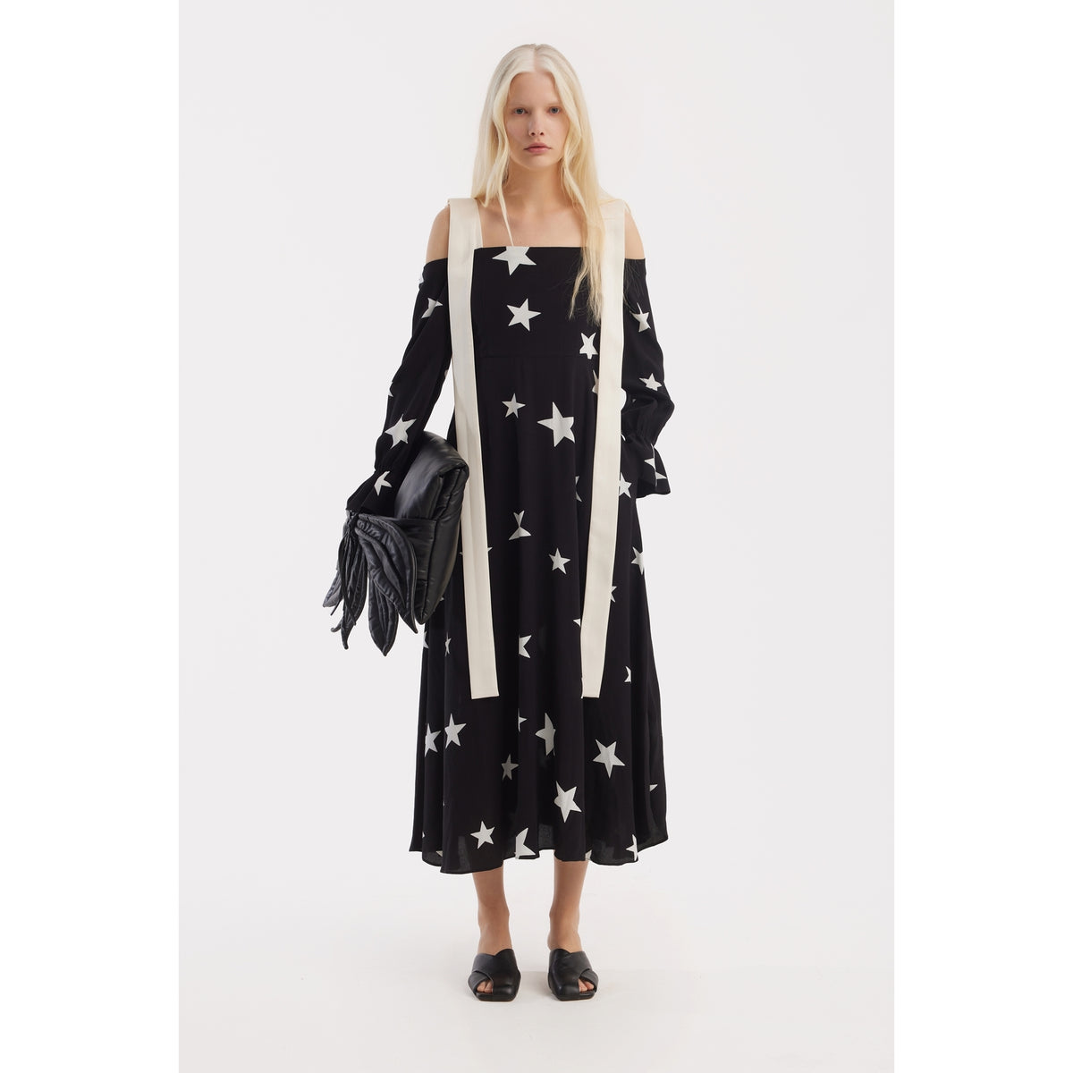 A woman with long blonde hair wears a JULIA ALLERT Off-Shoulder Midi Dress With Contrast Straps in black with star print, black slide sandals, a black fringed bag, and has a long light-colored scarf draped around her neck.
