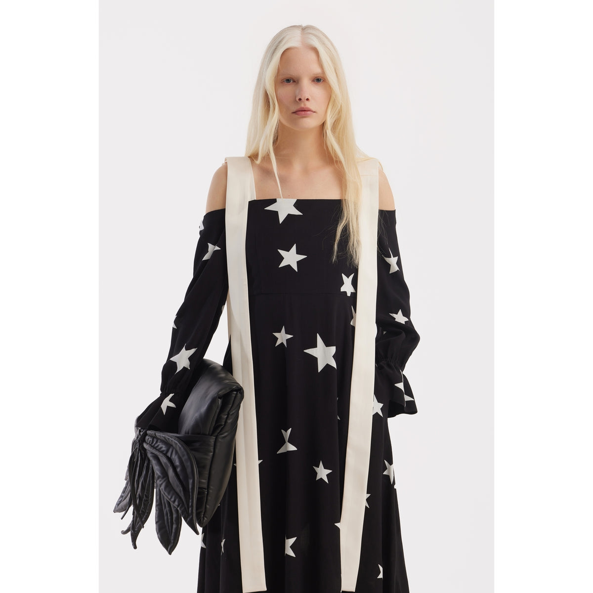 A person with long blonde hair wears the JULIA ALLERT Off-Shoulder Midi Dress With Contrast Straps in Black Print Stars, holding a black wing-shaped quilted accessory against a plain white background.