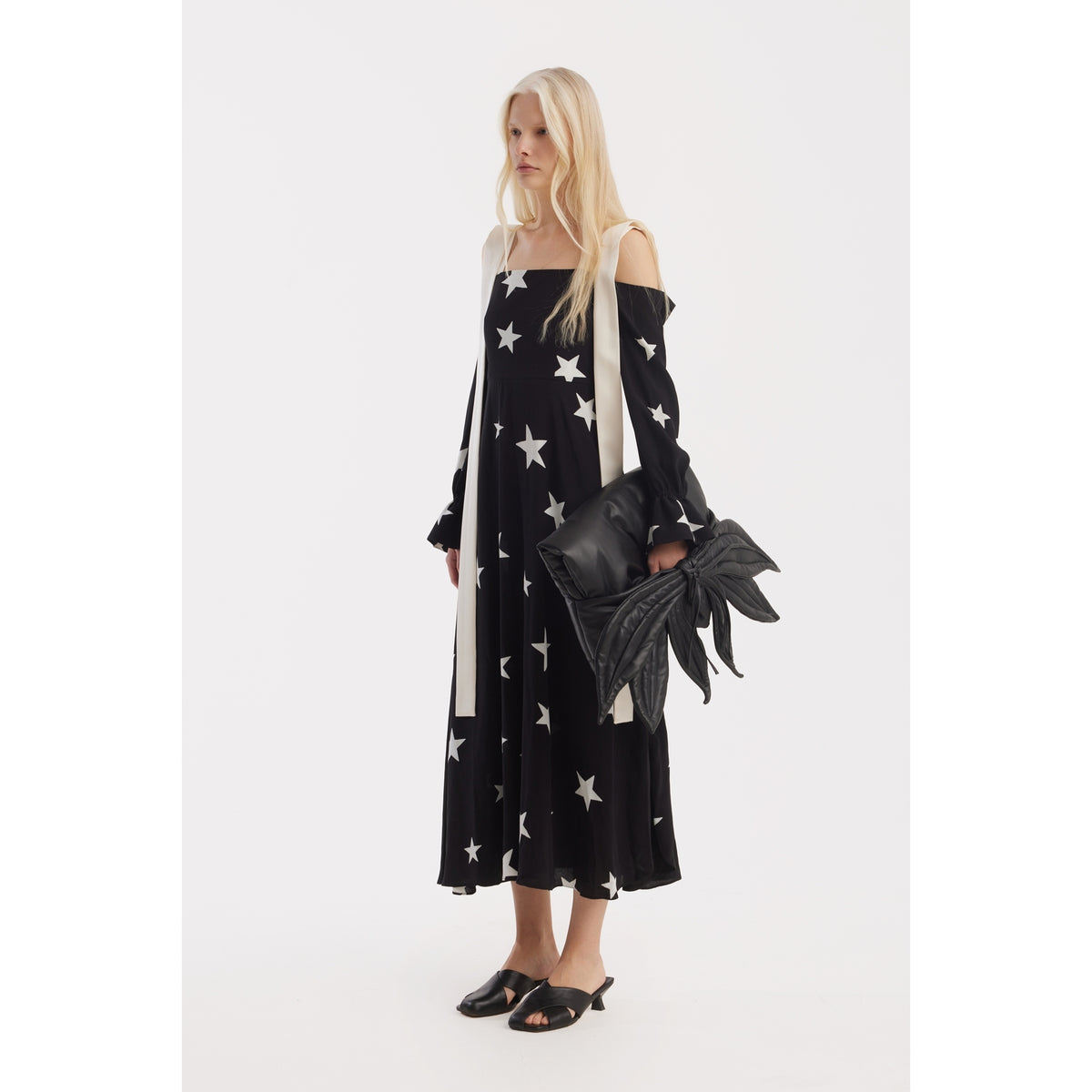 A woman with long blonde hair stands against a white background, wearing the JULIA ALLERT Off-Shoulder Midi Dress With Contrast Straps in black star print, paired with black slip-on shoes and a large sculptural shoulder bag with bow-like details.