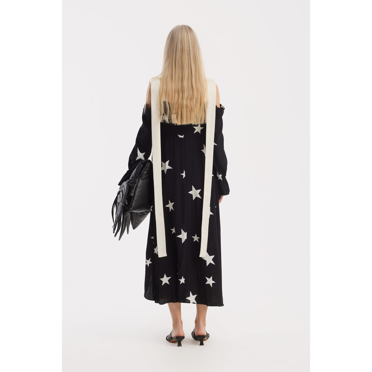 A woman with long blonde hair faces away, wearing the JULIA ALLERT Off-Shoulder Midi Dress With Contrast Straps in black print stars, styled with black heeled sandals and a sculptural black bag, set against a plain white background.