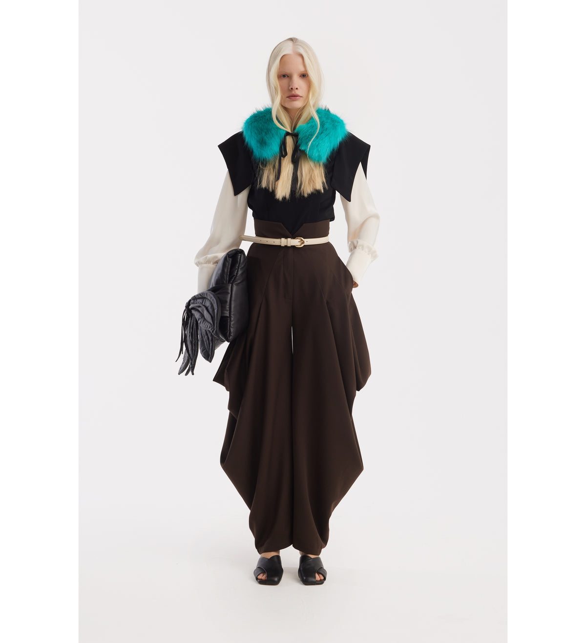 A woman stands against a white background, wearing a cream blouse, JULIA ALLERT Couture Draped Wide-Leg Pants in brown, a turquoise fur collar, black open-toe shoes, and holding a black textured handbag.
