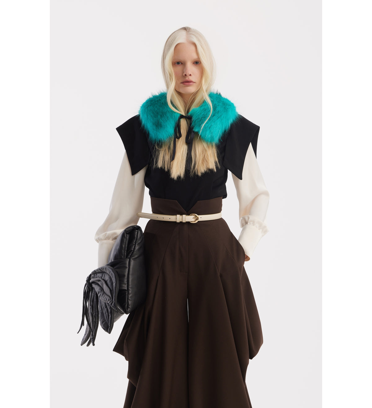 A person with long blonde hair wears a black and white top with a bright teal fur collar, JULIA ALLERT Couture Draped Wide-Leg Pants in brown, and holds a large black textured bag against a plain white background.