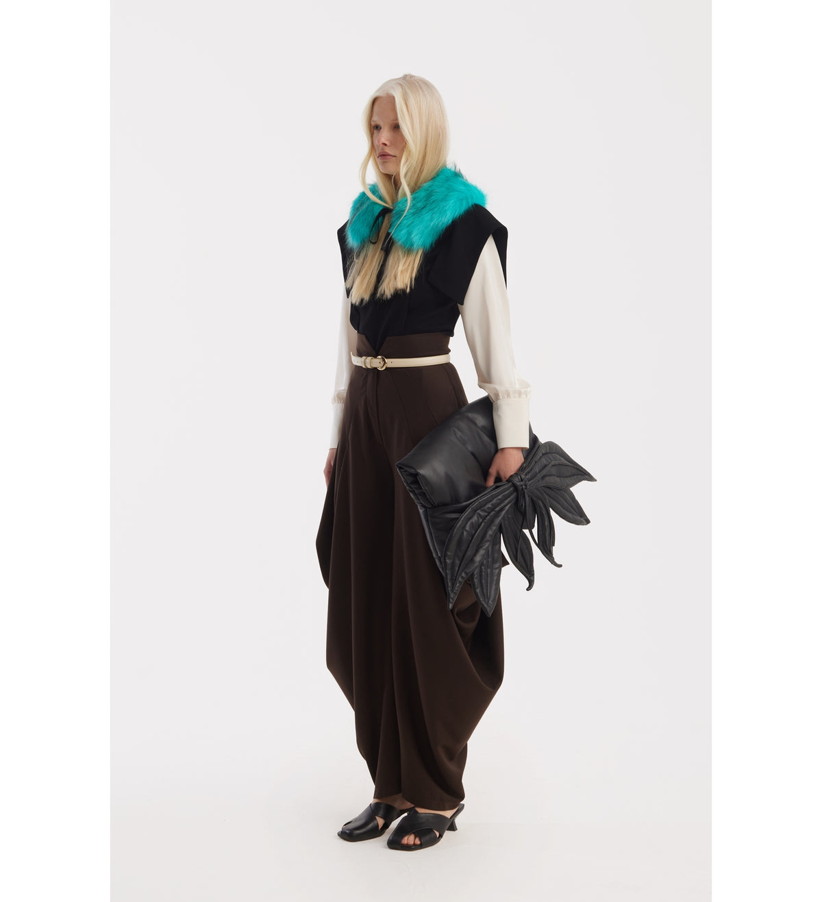 A person with long blonde hair wears a cream blouse, brown JULIA ALLERT Couture Draped Wide-Leg Pants, a black top with a teal fur collar, black sandals, and holds a large black leaf-shaped clutch against a plain white background.
