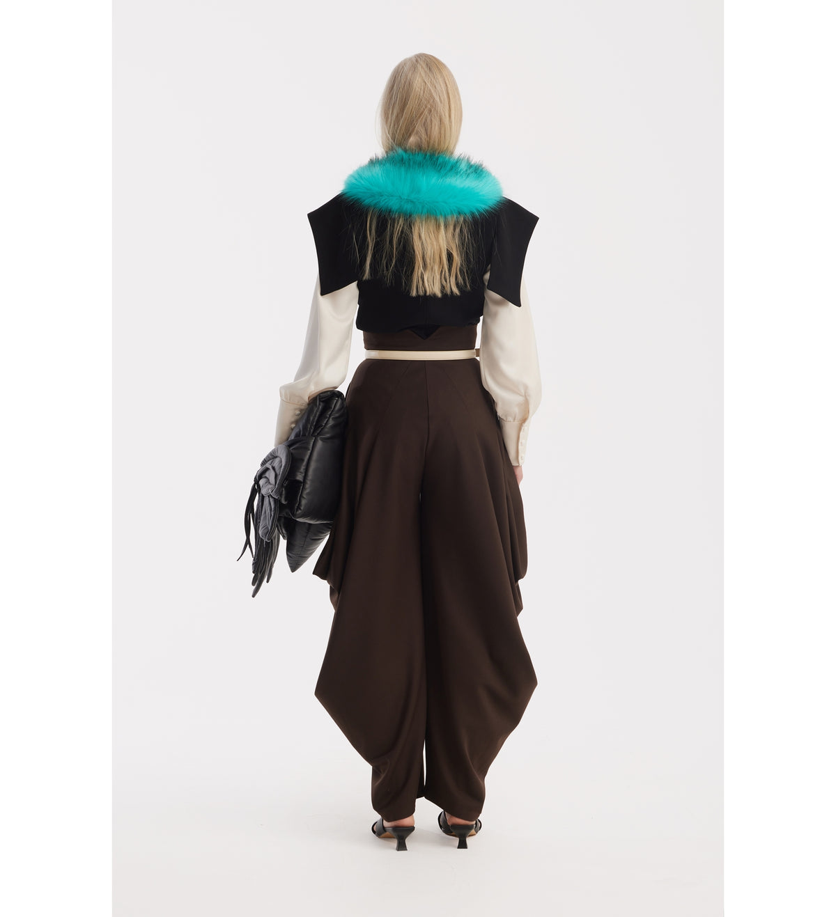 A person with long blonde hair, facing away, wears a cropped black top with a bright teal fur collar, JULIA ALLERT Couture Draped Wide-Leg Pants in brown, and black heels, holding a large black bag against a plain white background.