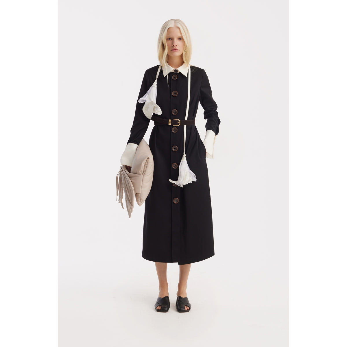 A blonde woman wears the JULIA ALLERT Monochrome Midi Shirt Dress - Black &amp; White with large buttons and a belt, holding a beige bag and gloves. Two white face masks hang from her neck against a plain white background.