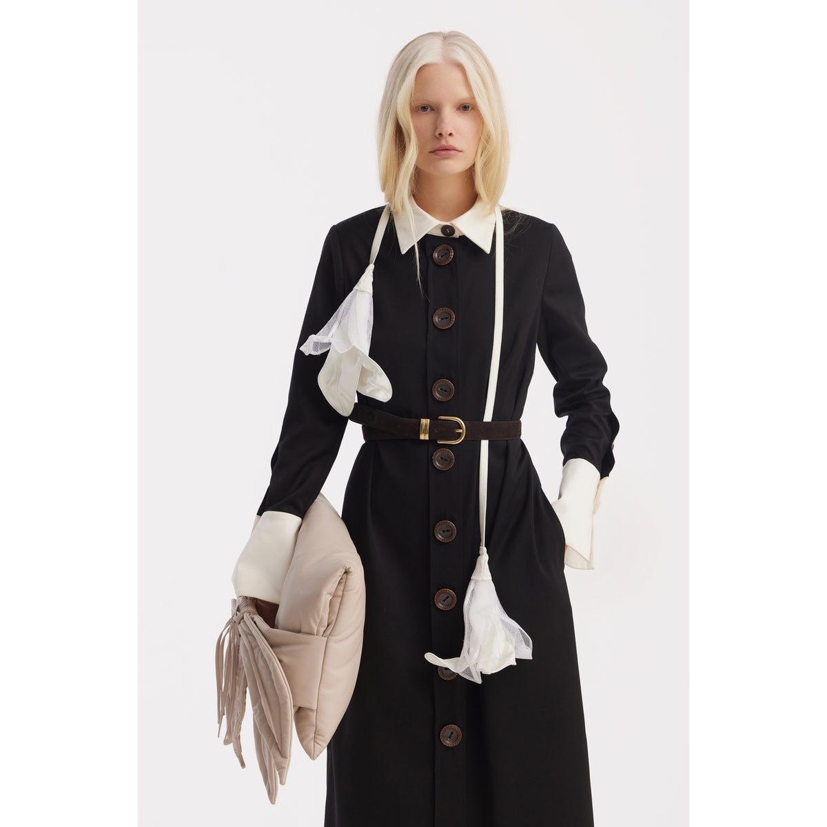 A woman with long blonde hair wears a black coat with white cuffs and collar, accessorized with a white scarf and holding the JULIA ALLERT Petal Faux Leather Evening Clutch Bag in Ecru against a plain white background.