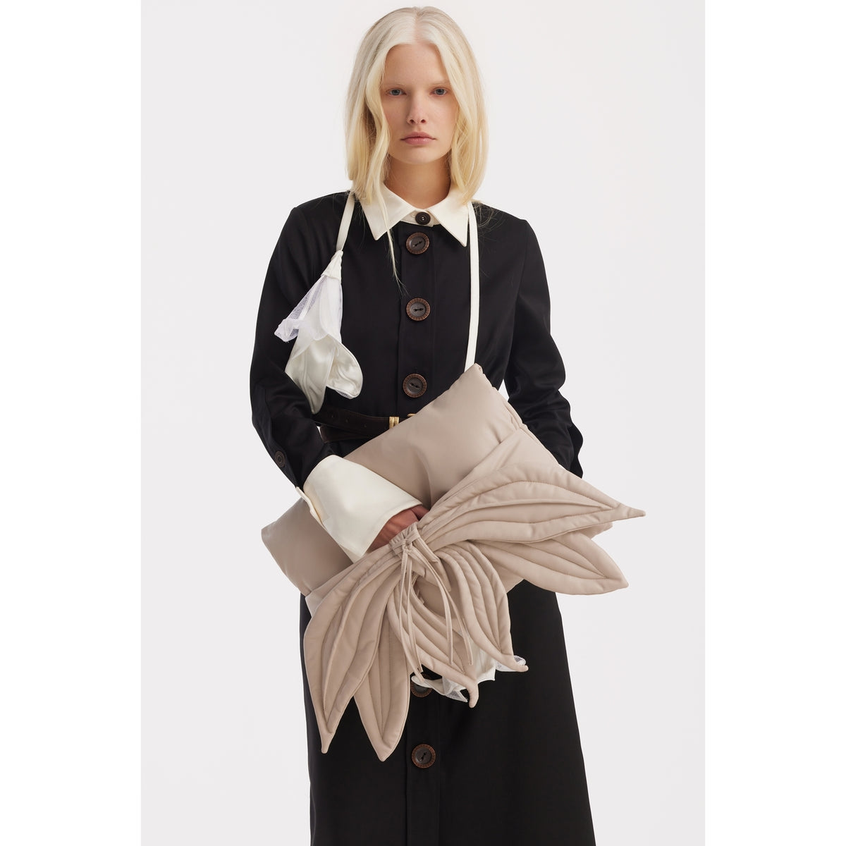A woman with long blonde hair wears a black trench coat with white cuffs and collar. She holds the JULIA ALLERT Petal Faux Leather Evening Clutch Bag in Ecru and a white eye mask, her expression neutral against a plain background.