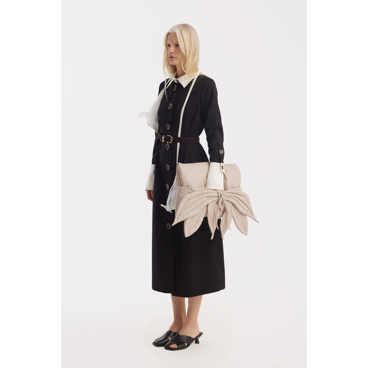 A woman stands against a white background wearing a long black buttoned dress, black sandals, and holding the JULIA ALLERT Petal Faux Leather Evening Clutch Bag in ecru with leaf-inspired details.