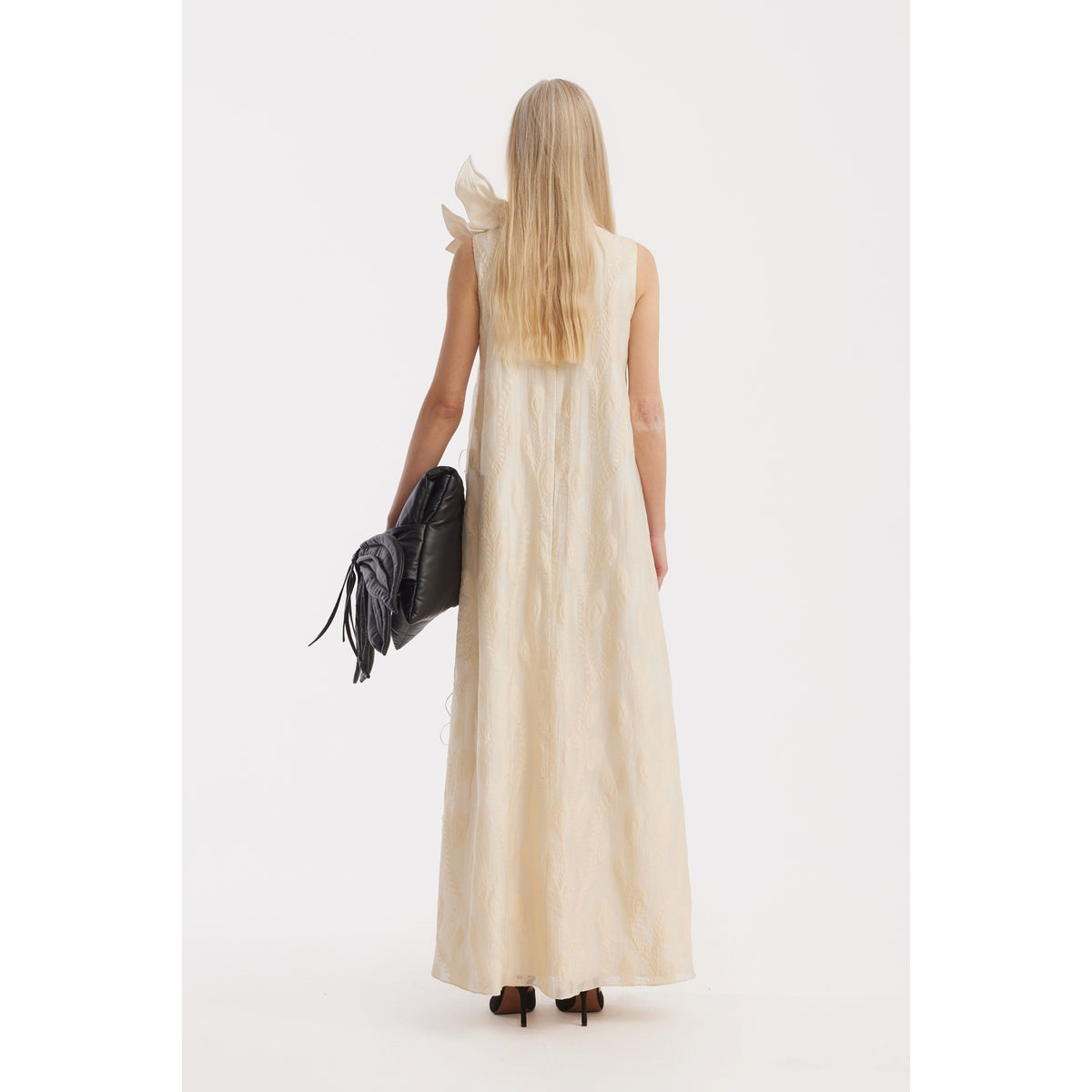 A woman with long blonde hair, seen from behind, wears the JULIA ALLERT Ivory A-Line Flower Maxi Dress—a sleeveless couture gown with a textured pattern and large shoulder bow. She holds a black clutch and wears black heels.