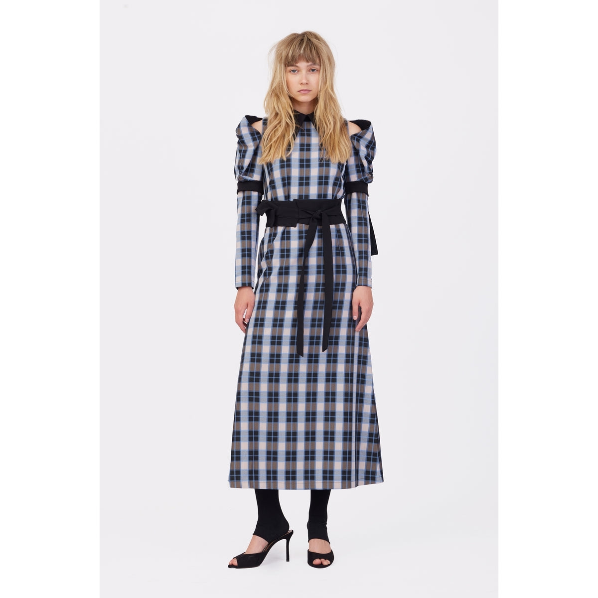 A woman with long, wavy blonde hair and a neutral expression models the JULIA ALLERT Stylish Cold Shoulder Long Sleeve Dress With Belt in Blue Check against a white background, paired with black tights and heeled sandals.