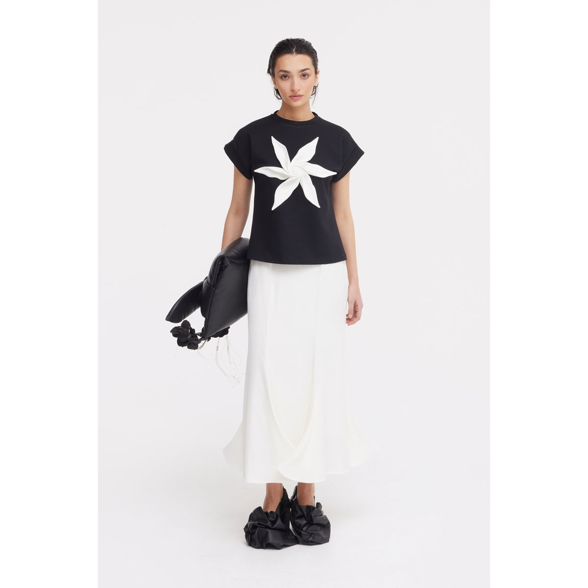 A woman stands against a white background wearing JULIA ALLERT’s black Crew-Neck Jersey T-Shirt With Flower, paired with a long white skirt, black oversized shoes, and holding a large black bag.
