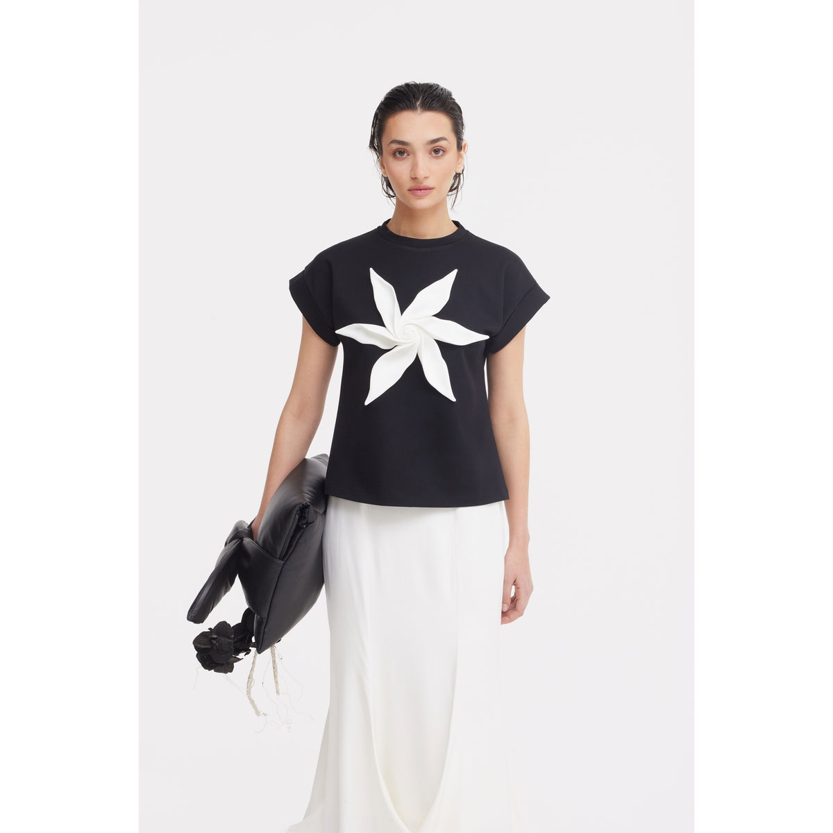A woman stands against a white background wearing the JULIA ALLERT Crew-Neck Jersey T-Shirt With Flower in black, paired with a long white skirt and holding a black handbag.