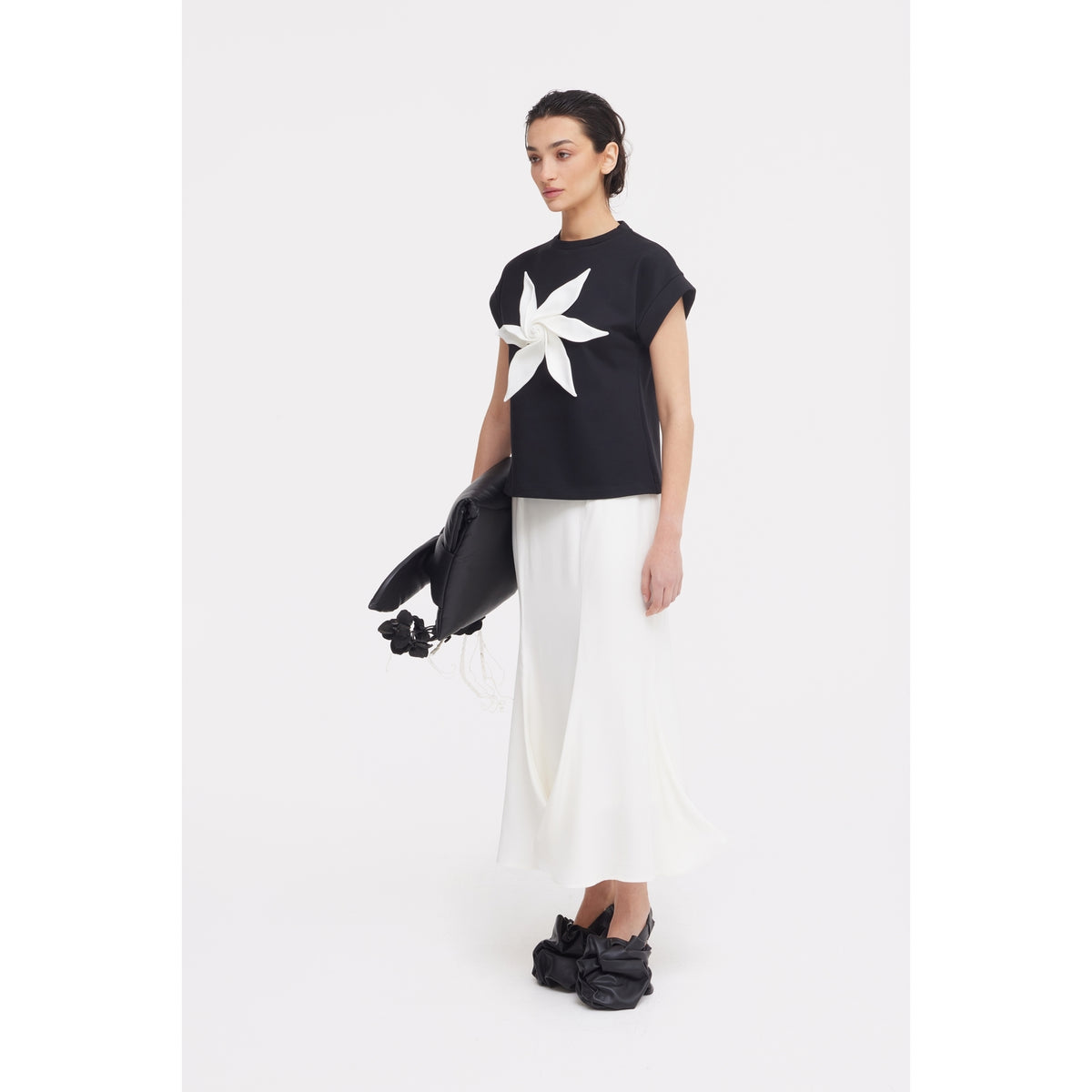 A woman stands against a white background wearing JULIA ALLERT's black Crew-Neck Jersey T-Shirt With Flower, a long white skirt, black bow-detailed shoes, and holding a large black bag.