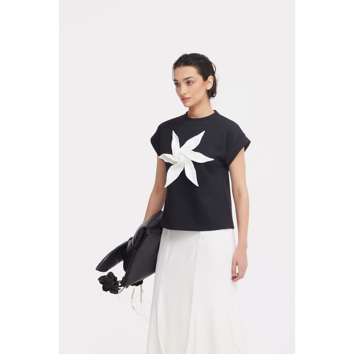 A woman wears the JULIA ALLERT Crew-Neck Jersey T-Shirt With Flower in black and a white skirt, standing against a light background and holding a black handbag with a floral detail in her left hand.