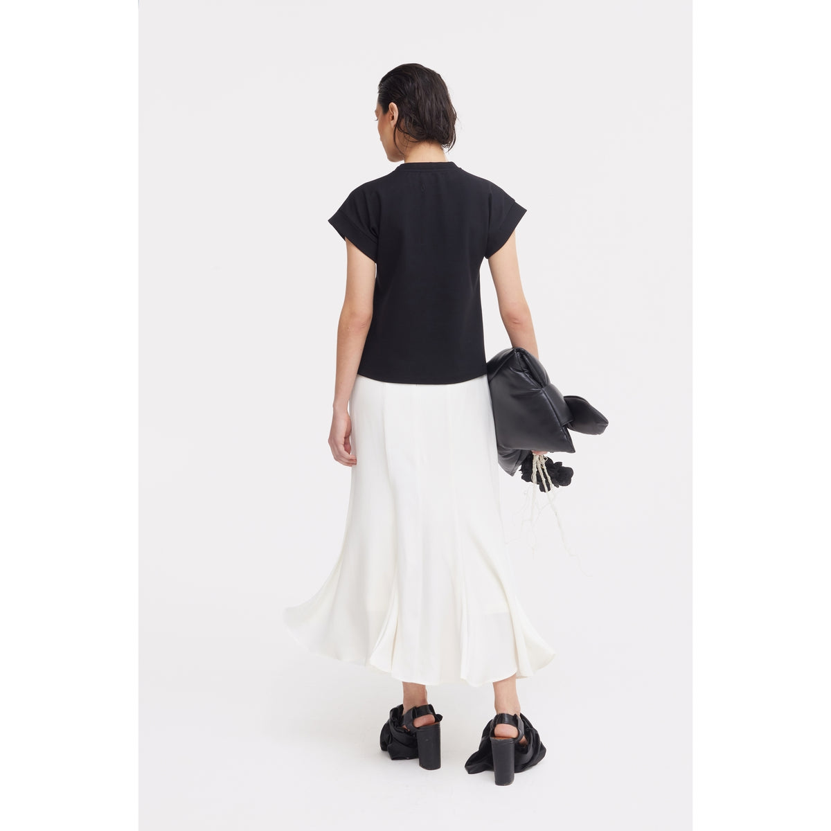 A person faces away against a white background, wearing the JULIA ALLERT Crew-Neck Jersey T-Shirt With Flower in black, paired with a flowing white skirt and black heeled sandals. They hold a black jacket and small black bag in their left hand.